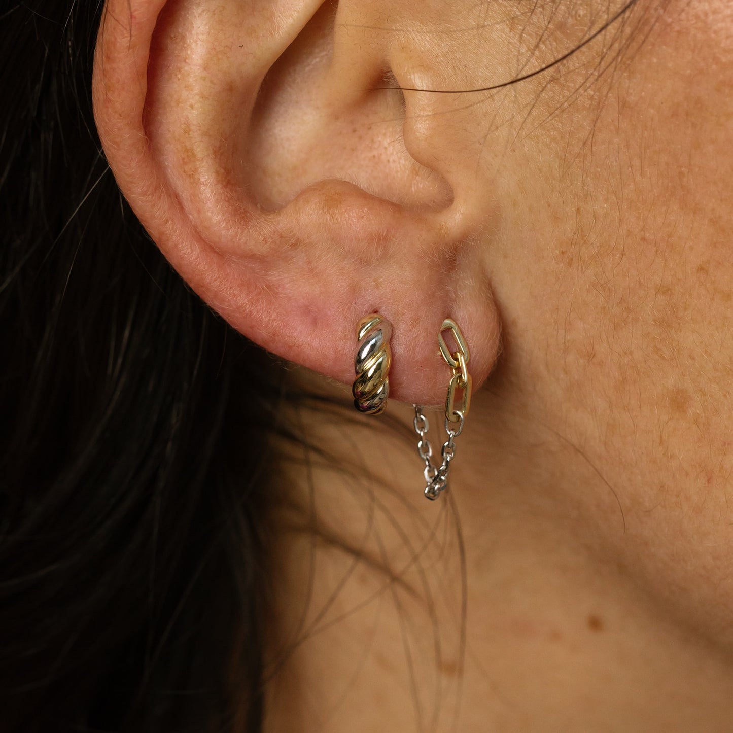 A close-up of a light-skinned person’s ear showcases the Solid Gold Mixed Metal Chain Dangle Huggies—a Local Eclectic exclusive—featuring twisted, chunky gold hoops and chain huggies connecting two mixed metal hoops. Brown hair is partly visible.