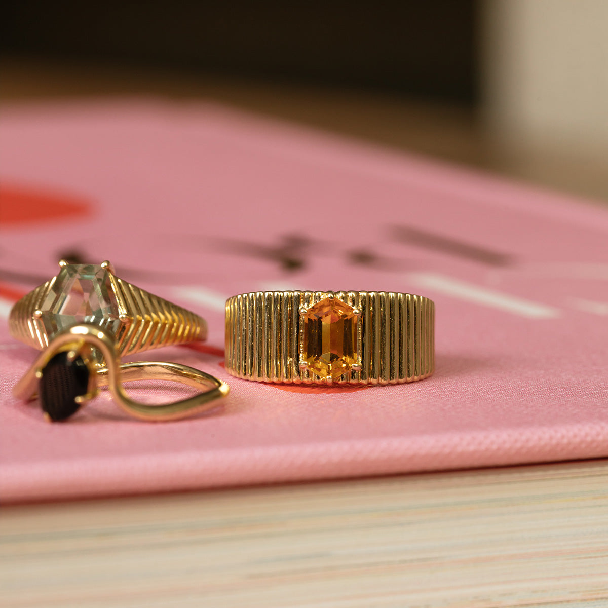 Two gold rings rest on a pink surface; one is the Solid Gold Citrine Bold Fluted Cigar Band with a rectangular citrine, while the other features a clear stone in a twisted setting.