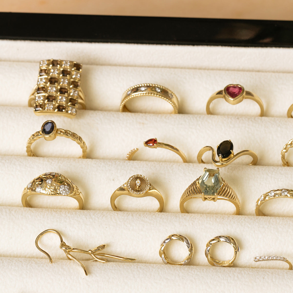 A cream-colored velvet tray displays a Solid Gold Garnet and Sapphire Open Front Band, along with multiple 9k yellow gold rings, gold earrings, and small gold hoop earrings.