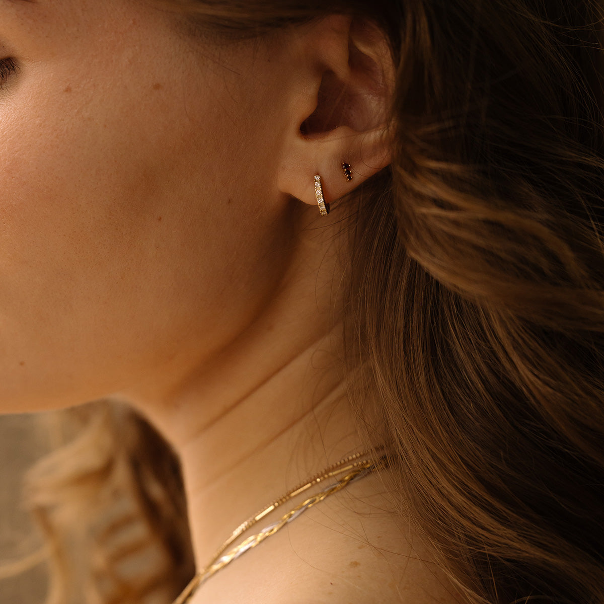 A woman wears Solid Gold Diamond Huggie Hoops and a stud in her ear, with wavy brown hair flowing past and layered gold necklaces. Her softly lit skin highlights the sparkle of the huggie hoops.