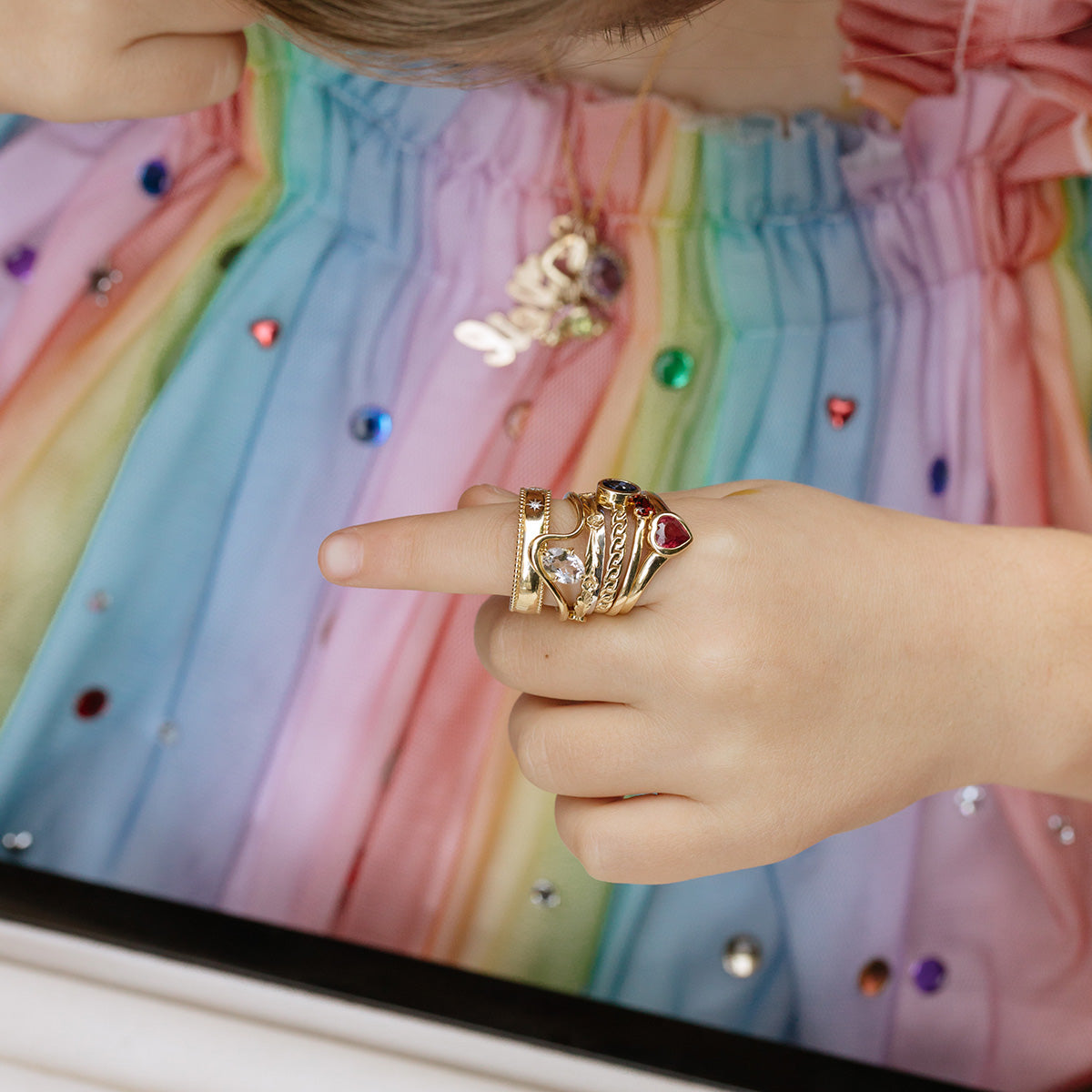 A child wearing a rainbow-colored dress with rhinestones displays gold rings, including the Solid Gold Mixed Metal Filigree Band, on their finger. The child also has a name pendant necklace.