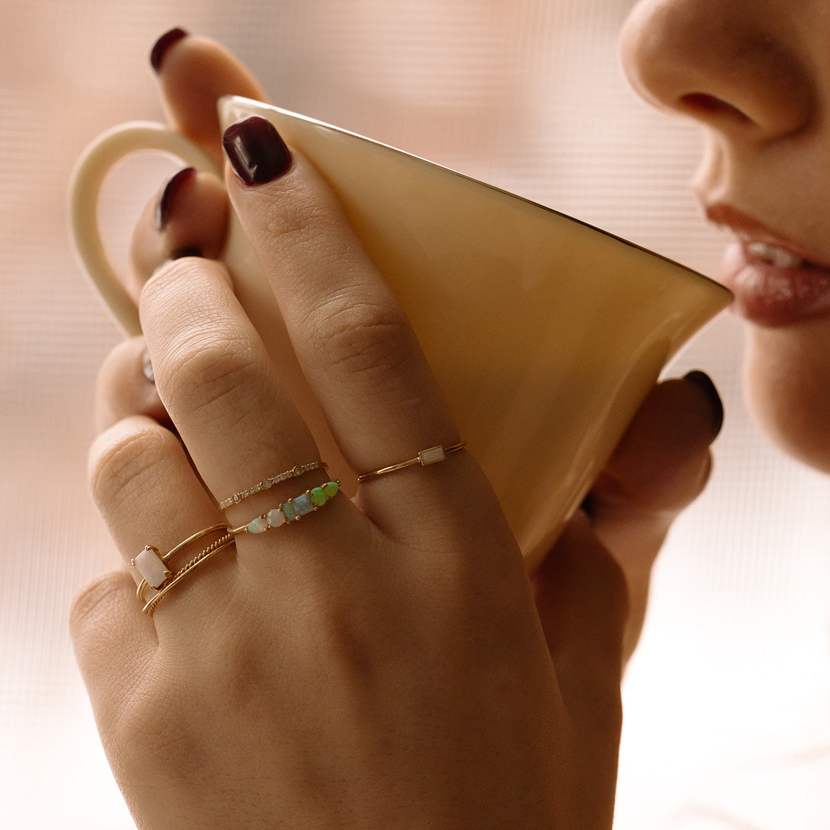 A close-up of a woman with dark nail polish holding a beige mug near her lips, wearing several delicate gold bands and gemstone rings, including the Solid Gold Crossover Ring Set.
