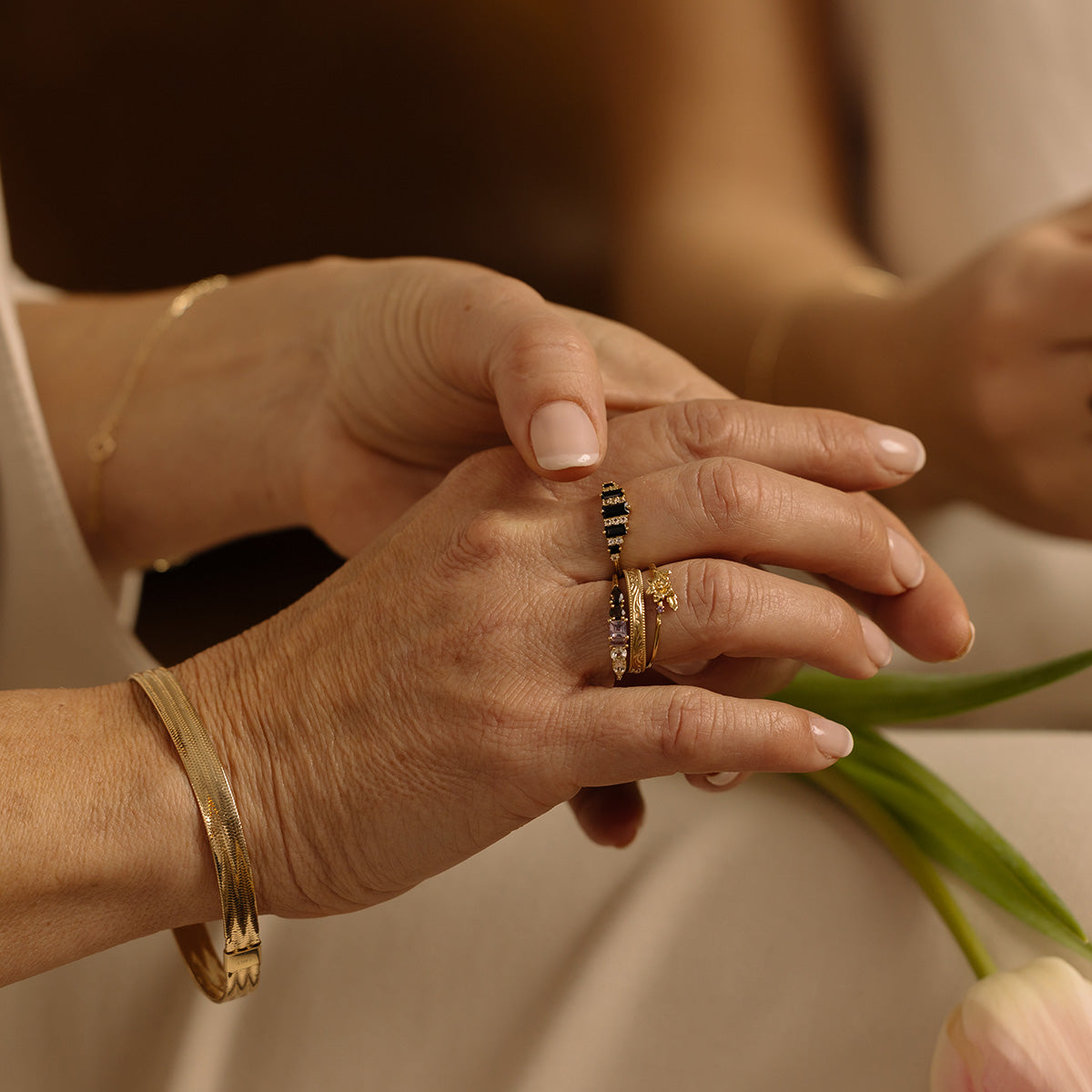 A close-up of two hands, one gently holding the other, both adorned with gold jewelry including a vintage-inspired Solid Gold Filigree Ring and bracelets. A tulip appears in the background, enhancing the warm, tender mood.