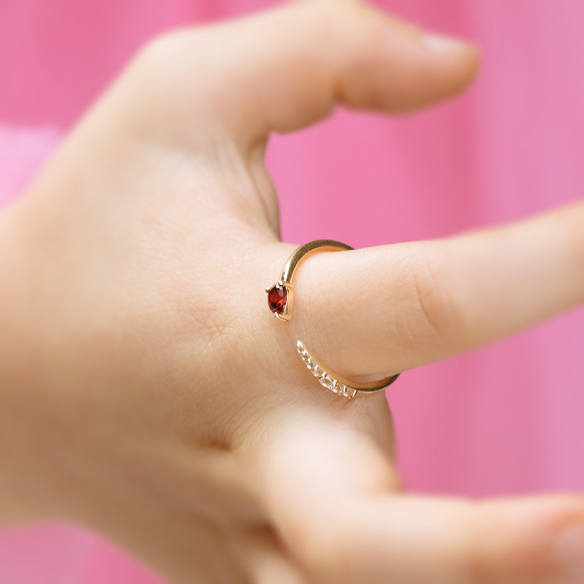A close-up of a hand wearing the Solid Gold Garnet and Sapphire Open Front Band on the middle finger, its vibrant gems shining against a blurred pink background.