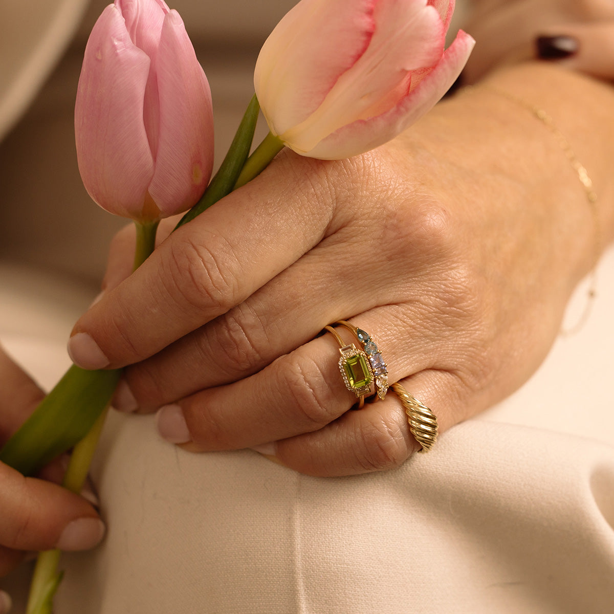 A close-up of a hand holding two pink tulips, adorned with Family Gold rings—one is the Solid Gold Vintage Inspired East West Peridot Ring with diamonds, the other a vintage charm ring with a unique twisted design. The background is softly blurred.