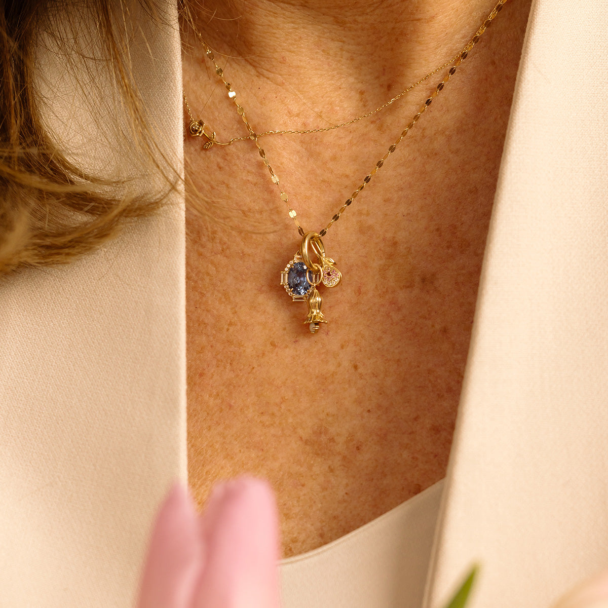 A close-up of a woman wearing layered gold necklaces with gemstone pendants and the Solid Gold Foodie Charm, including a blue stone, over a cream blouse. A blurry pink flower is visible in the foreground.