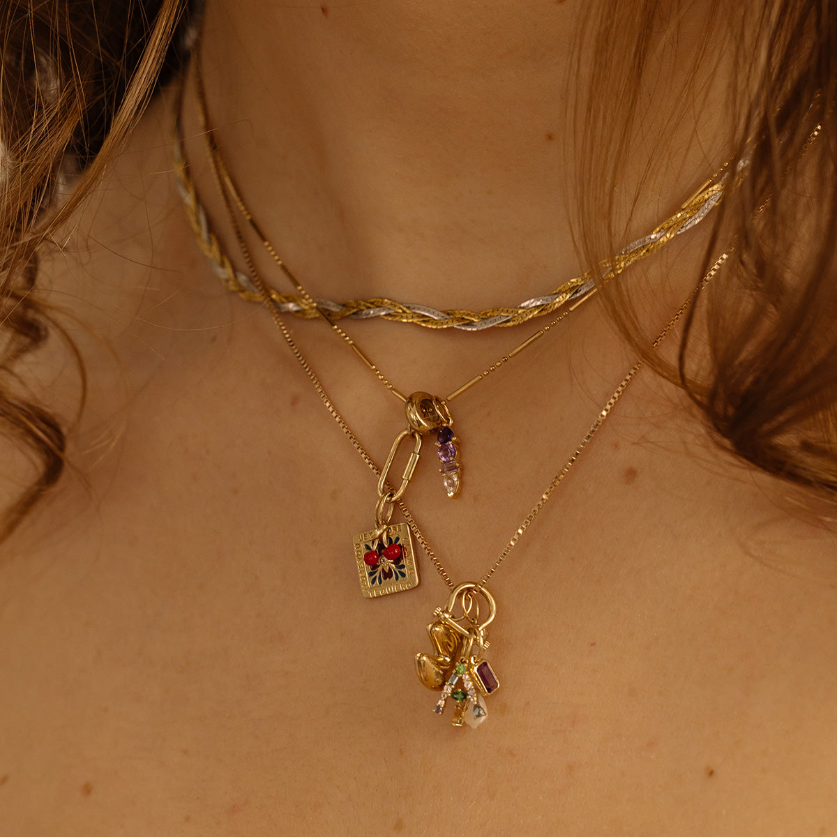 Close-up of a womans neck adorned with three layered necklaces: a twisted choker, a gold chain featuring the Solid Gold Gemstone Letter Charm with a purple bead, and two gold chains embellished with colorful pendants and charms.
