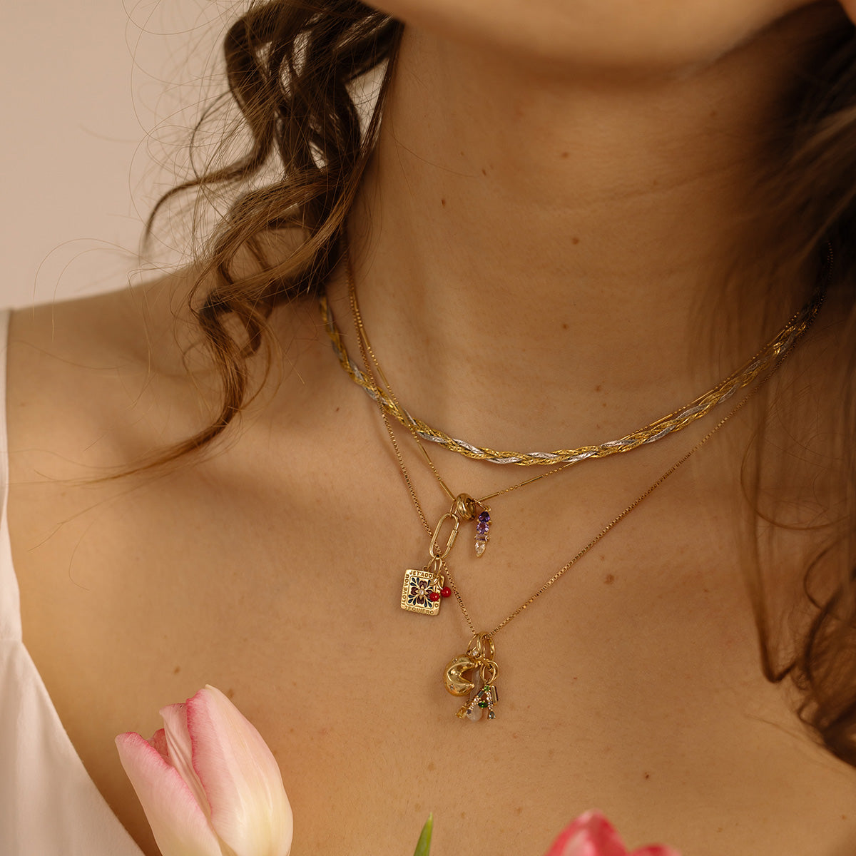 A woman with curly hair wears layered gold necklaces featuring the Solid Gold Body Language Charm, a Local Eclectic exclusive, paired with a white top and holding pink tulips near her chest.