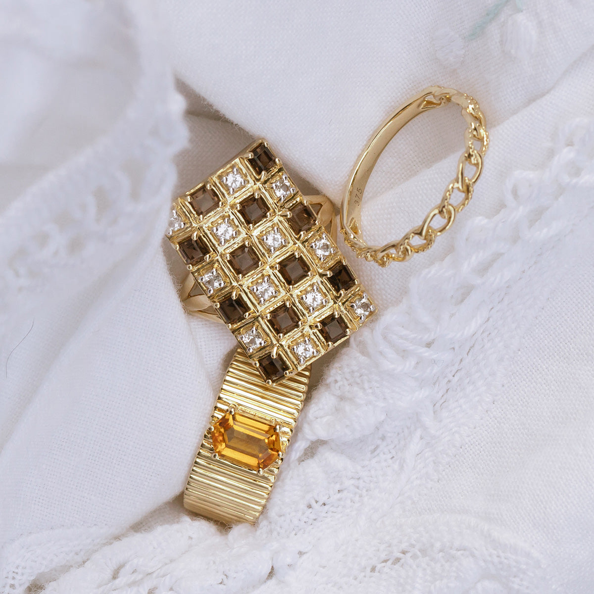 Three gold rings rest on white embroidered fabric: one is the Solid Gold Checkerboard Gemstone Shield Ring, another features a yellow rectangular stone reminiscent of smoky quartz, and the third is a delicate chain-style band.