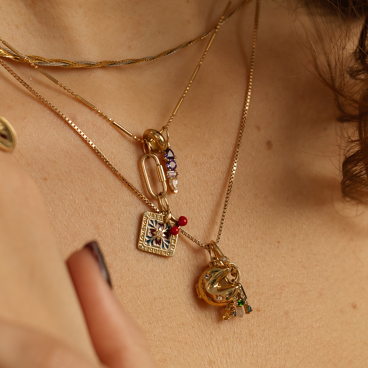 A close-up of a person wearing multiple gold necklaces, including the Solid Gold Statement Charm with colorful beads, a 9k solid yellow gold square, and a pearl and blue topaz pendant. The persons hand is partially visible.