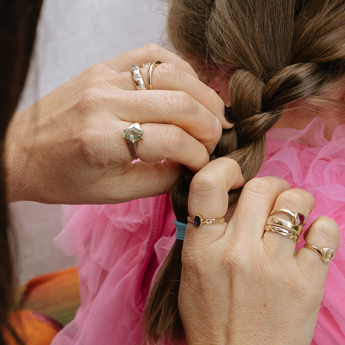 Close-up of hands wearing a Solid Gold Blue Sapphire Chain Link Ring braiding a young girls brown hair, secured with a blue hair tie. The girl is in a ruffled pink top.