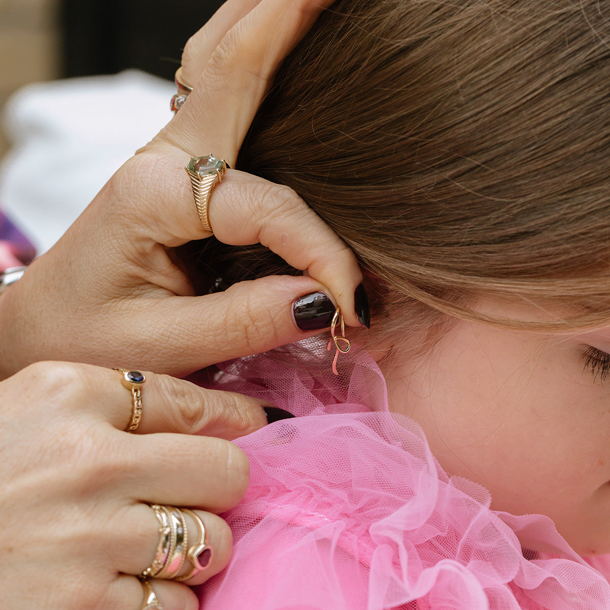 A close-up of an adult adjusting a young girls earring. The girl, in a pink ruffled top, has her brown hair gently tucked behind her ear by hands wearing the Solid Gold Blue Sapphire Chain Link Ring and several bracelets.