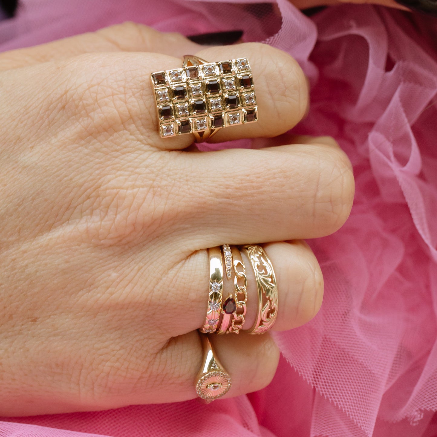 A hand wearing the Solid Gold Garnet and Sapphire Open Front Band along with three other ornate 9k yellow gold rings rests on soft pink tulle fabric.