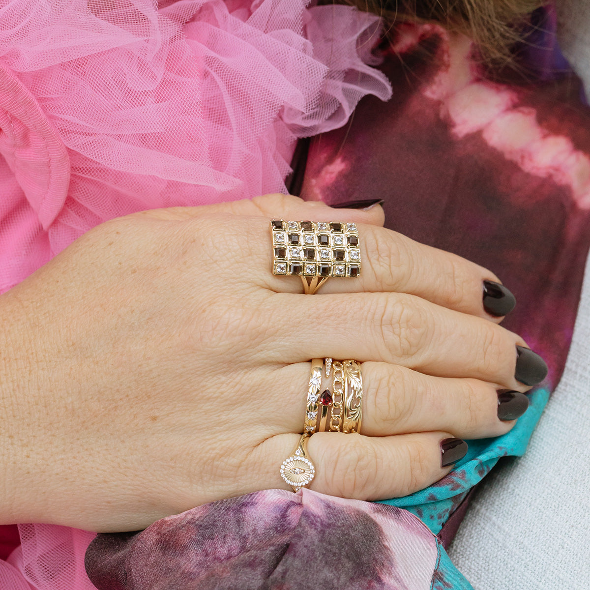 A close-up of a hand with dark-painted nails wearing the Solid Gold White Topaz Etched Signet Pinky Ring, surrounded by other gold bands, set against a vibrant, textured fabric background.