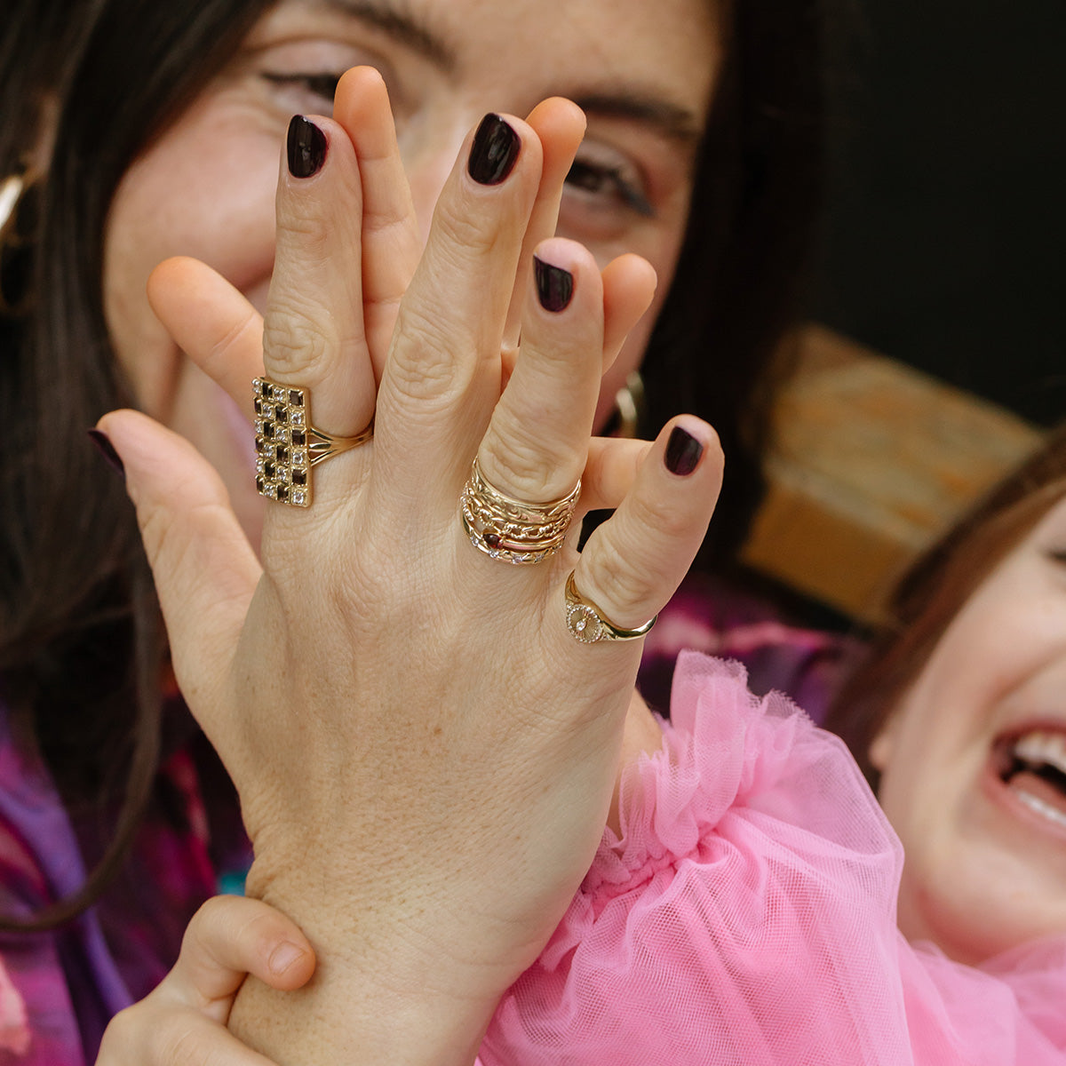 A close-up of two hands with dark nail polish, both wearing the Solid Gold Textured Chain Link Ring, pressed together in a high-five. In the background, two smiling people—one in a pink sleeve—are partially visible and laughing.