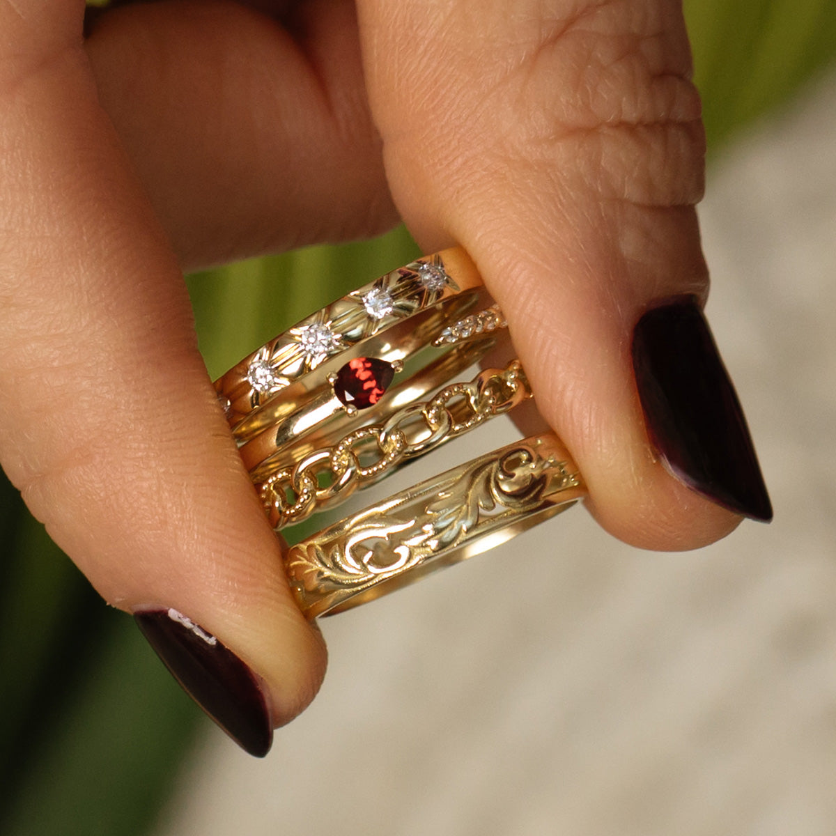 A hand with dark, glossy nail polish holds four gold rings: a Solid Gold Vintage Inspired Diamond Band, a chain ring, one with a red gemstone, and one engraved with swirls.
