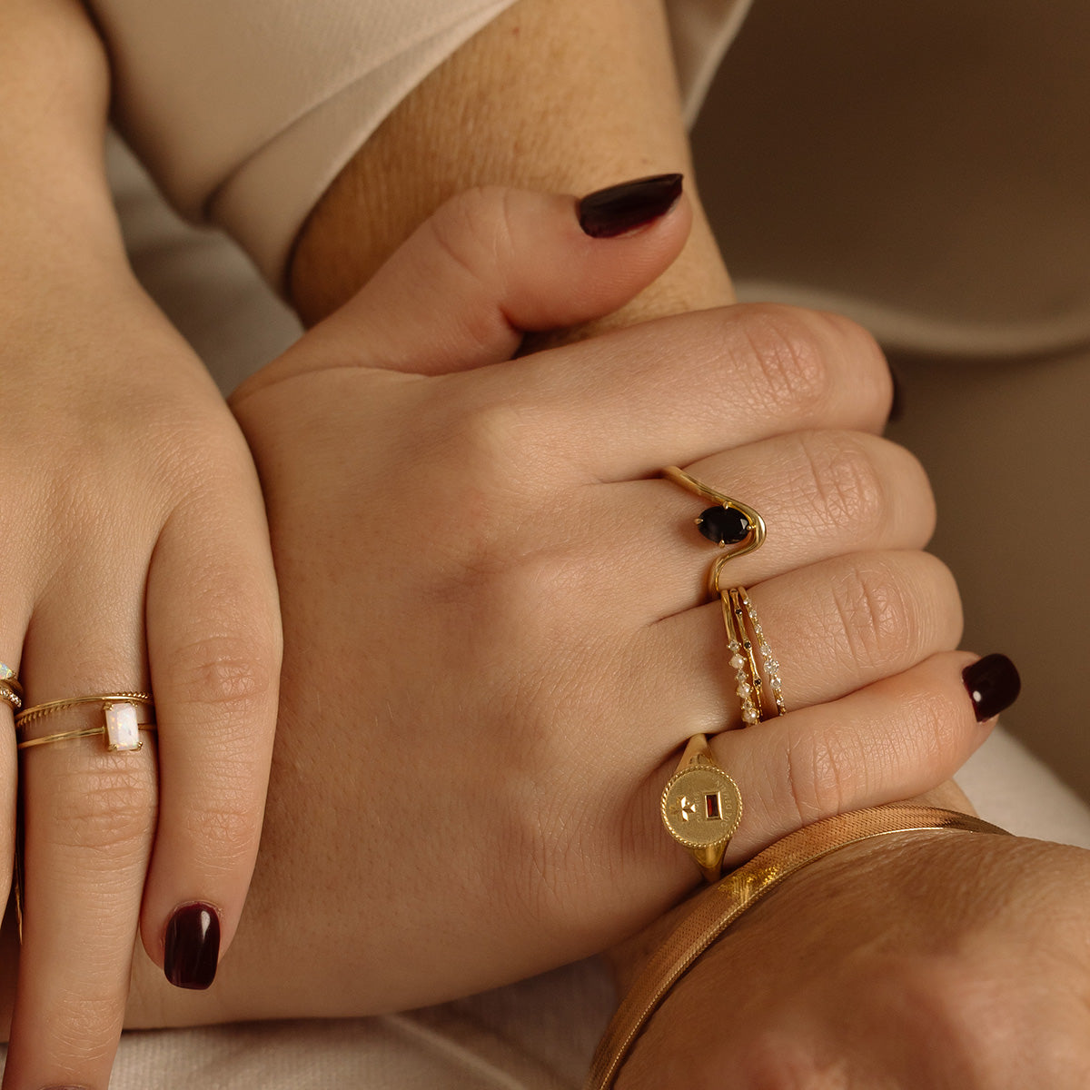 Two hands with dark burgundy nail polish, adorned with gold rings—including a Solid Gold Textured Diamond Band—a medallion, and a bracelet—are gently holding each other against a beige background.