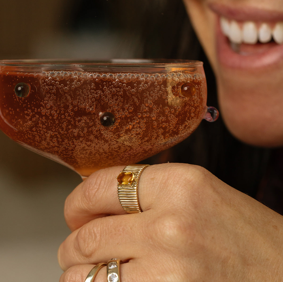 A smiling person holds a coupe of bubbly brown drink. Their partially visible face shows white teeth, and they wear the Solid Gold Citrine Bold Fluted Cigar Band, crafted in 9k solid yellow gold.