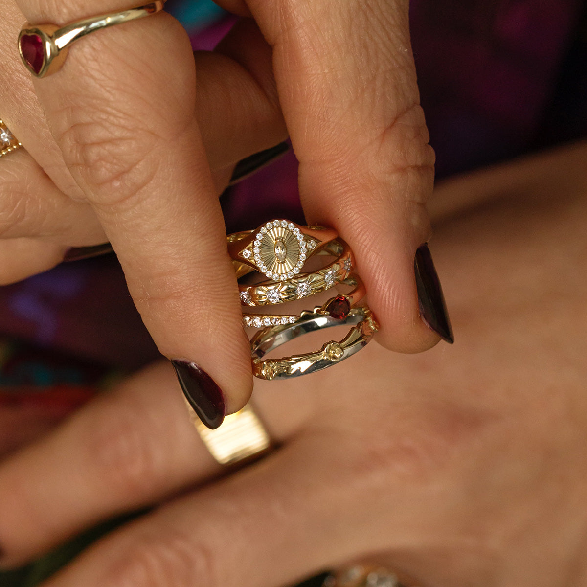 A hand with dark nail polish wears stacked gold rings, including the Solid Gold White Topaz Etched Signet Pinky Ring and gemstone pieces. Another hand in the background is adorned with a solid yellow gold ring.