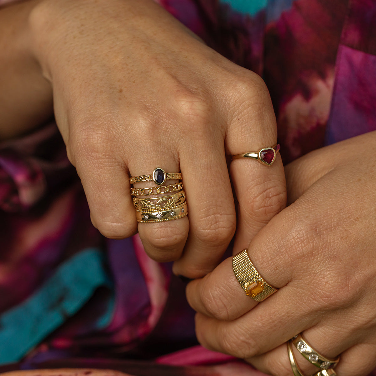 Close-up of two hands wearing gold rings, including the Solid Gold Openwork Filigree Ring with intricate detailing. The wearer is dressed in a vibrant purple and pink patterned garment. Rings showcase unique designs like heart-shaped stones and filigree work.