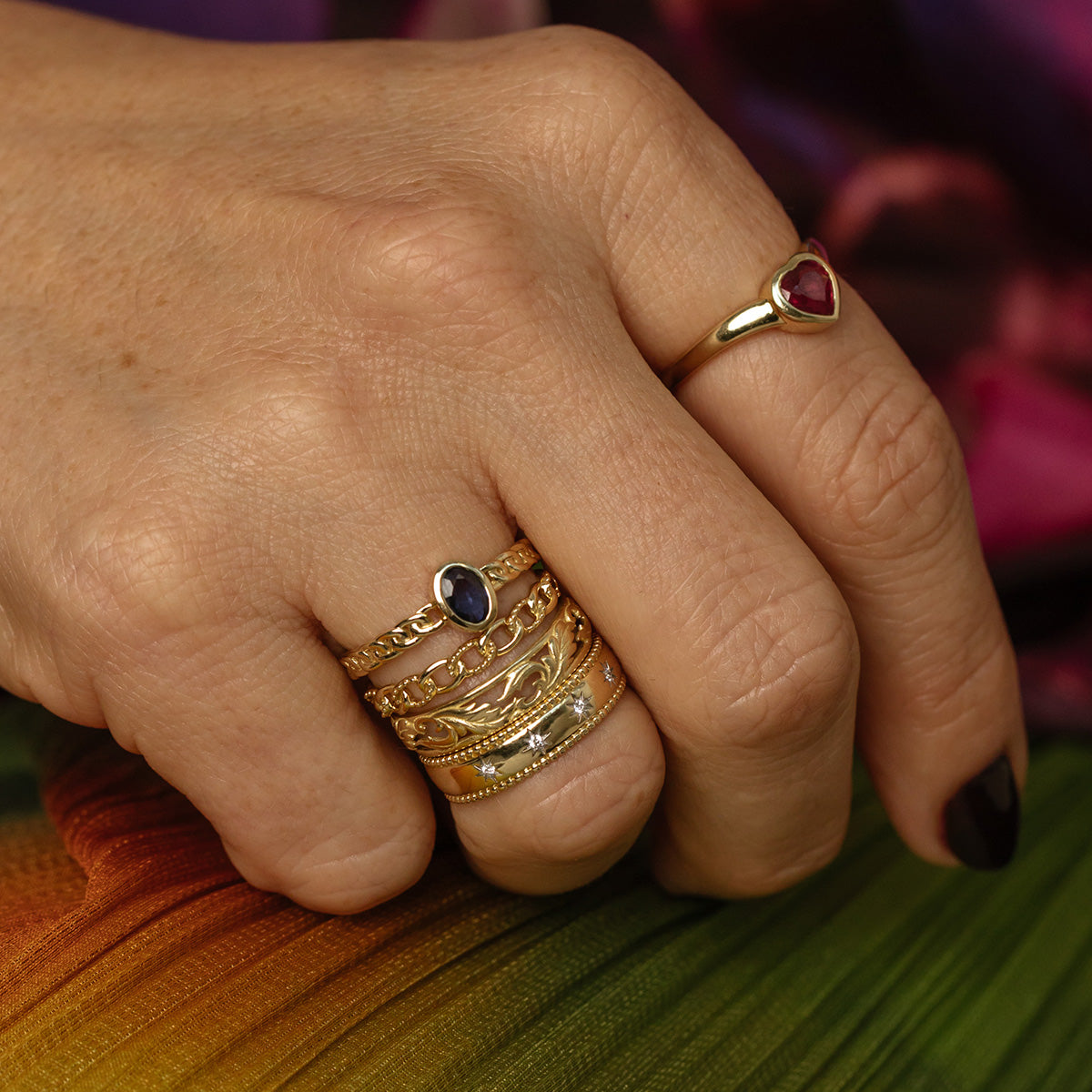 A hand with dark nail polish wears a Solid Gold Blue Sapphire Chain Link Ring, along with other gold rings, and rests on vibrant textured fabric.