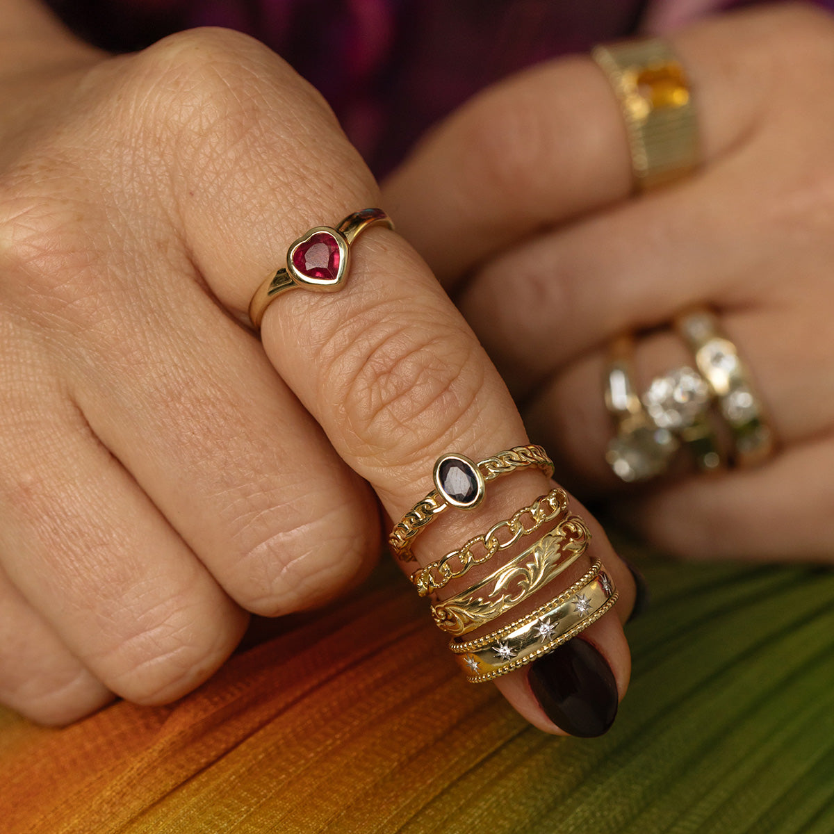 A close-up of two hands adorned with gold rings, including the Solid Gold Blue Sapphire Chain Link Ring, set against a rainbow-patterned fabric background. The rings display gemstones and intricate engravings.