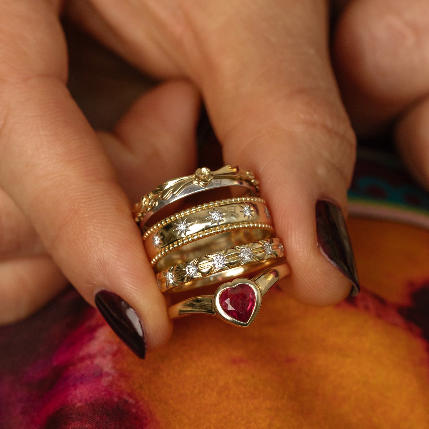A hand with dark nail polish holds several stacked gold rings, including the Solid Gold Mixed Metal Filigree Band, a two-toned band, and other yellow and white gold rings with decorative patterns and gemstones.