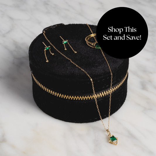 The Solid Gold 2025 May Capsule Emerald Full Set with Free Gift—including necklace, earrings, and ring—is displayed in a black velvet case with gold zipper. A black label reads, “Shop This Set and Save!”.