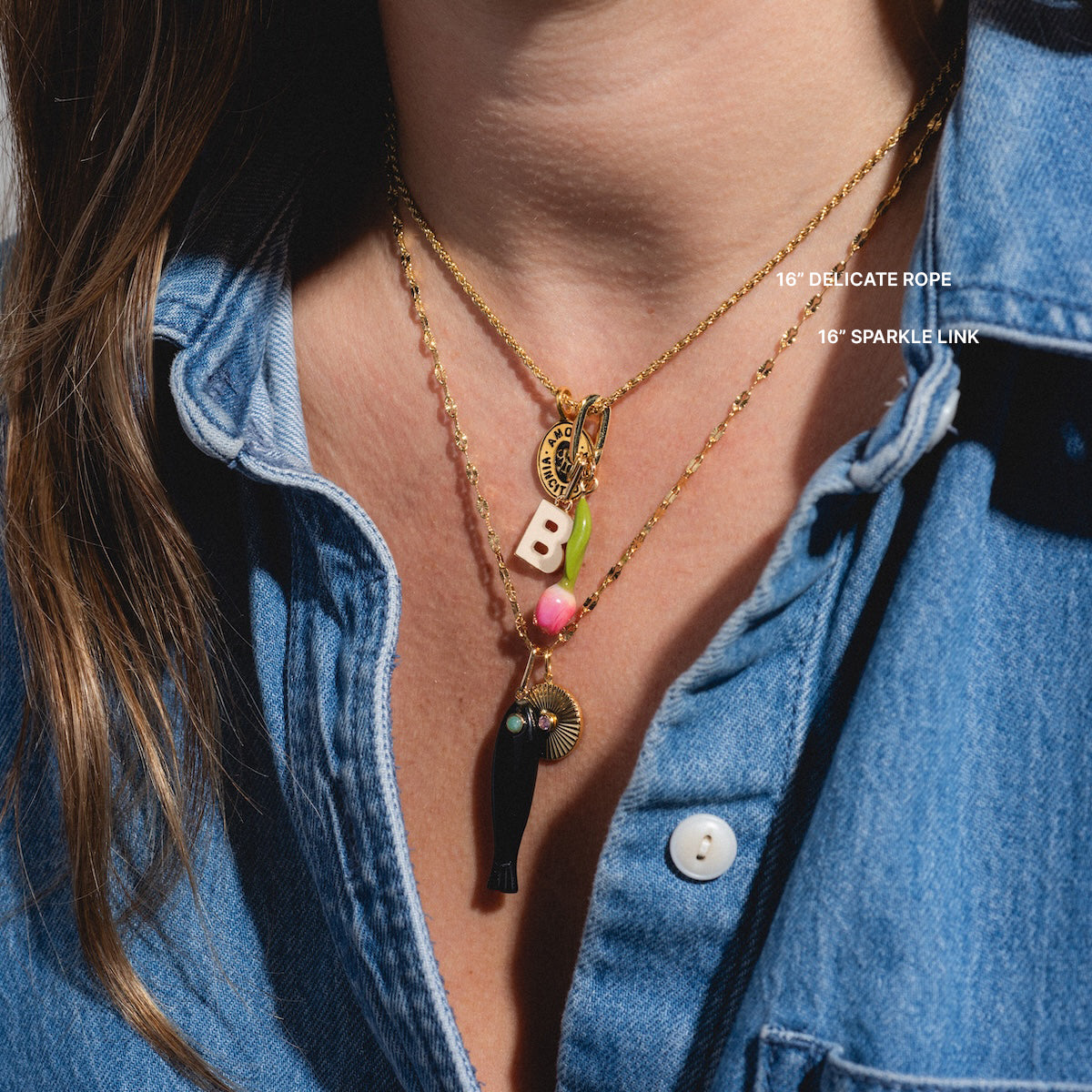 Someone wears a blue denim shirt and layered gold necklaces, including the Chain For Charms: a 14k gold plated chain featuring charms like the letter B, colorful beads, coin, fish, and fan. Labels show 16 DELICATE ROPE and 16 SPARKLE LINK styles.