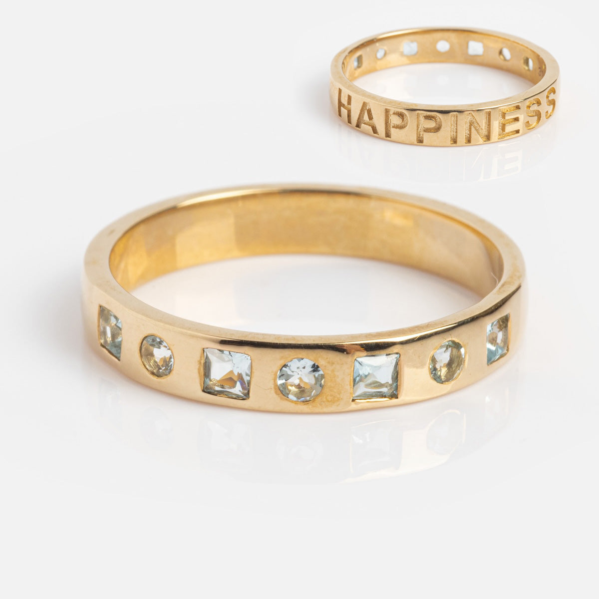 Two 9k solid yellow gold rings rest on a white background: one is engraved with HAPPINESS, while the Solid Gold Within You Birthstone Band Sample Size 7 features alternating round and square clear gemstones around its 3.1 mm band.