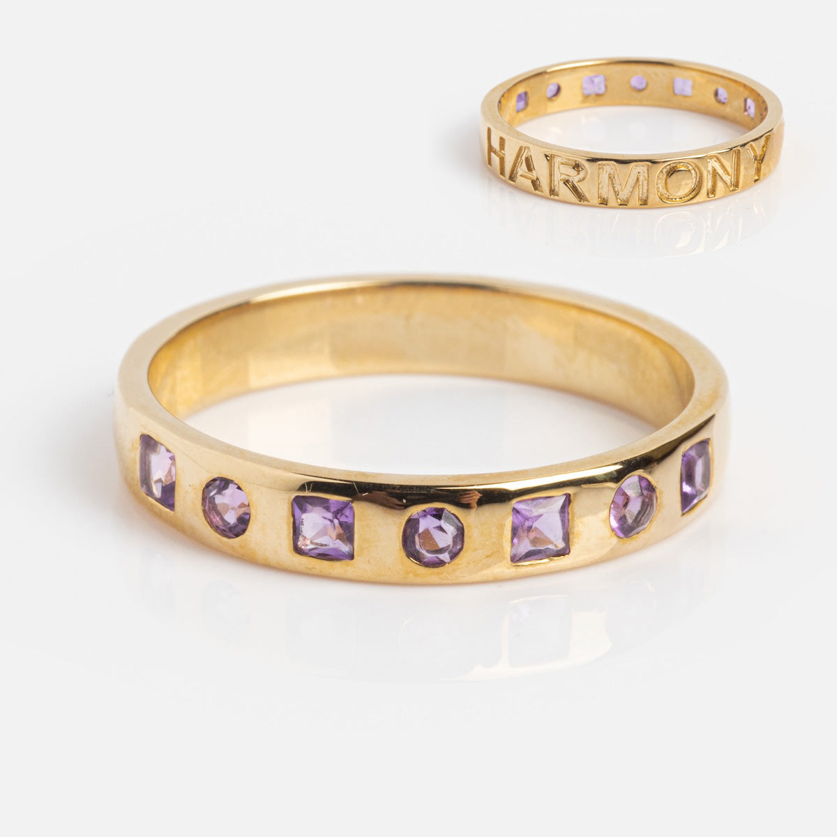 The Solid Gold Within You Birthstone Band Sample Size 7, a gold ring set with seven natural stones, is displayed on a white background alongside a 9k yellow gold HARMONY ring with purple stones.