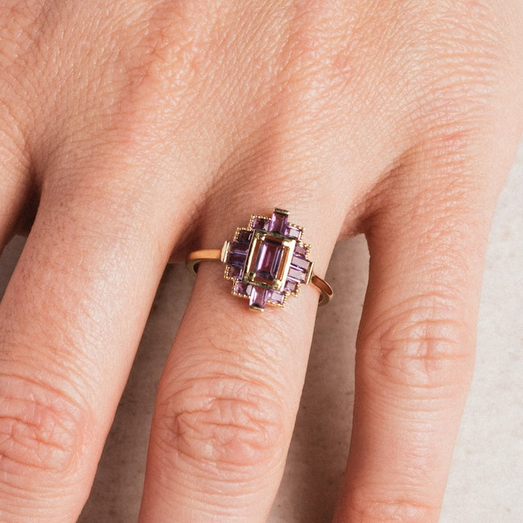 A hand wears the Solid Gold 2024 February Capsule Vintage Inspired Amethyst Statement Ring (Sample Size 9), featuring geometric rectangles of Amethyst set in yellow gold, displayed on the middle finger against a light background.
