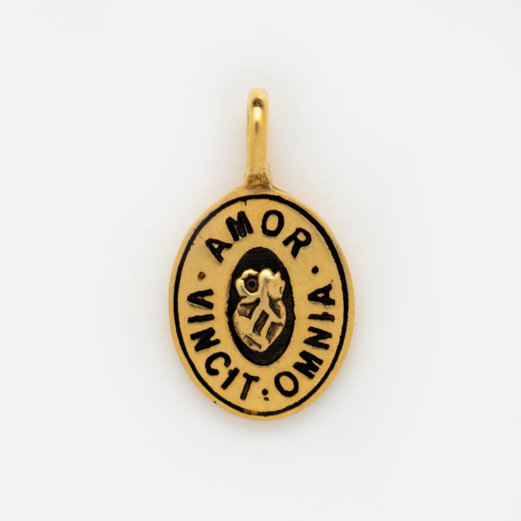 Charms by Hunt of Hounds features a gold vermeil oval pendant with a raised anatomical heart and AMOR VINCIT OMNIA engraved on a black background—an elegant symbol of timeless romance.