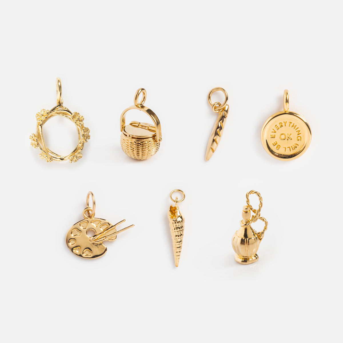 Seven small Charms by I Like It Here Club, each plated in 12k yellow gold, are displayed on a white background: a wreath, picnic basket, pea pod, engraved coin, artist palette with brushes, carrot, and an ornate pitcher.