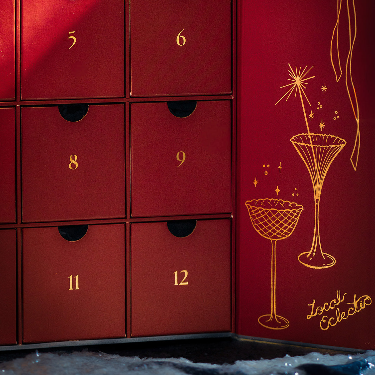 A close-up of the 2025 Jewelry Advent Calendar features red panels with gold numbers, doors numbered 5 to 12, gold cocktail glass illustrations, and "Local Eclectic" on the inside panel.
