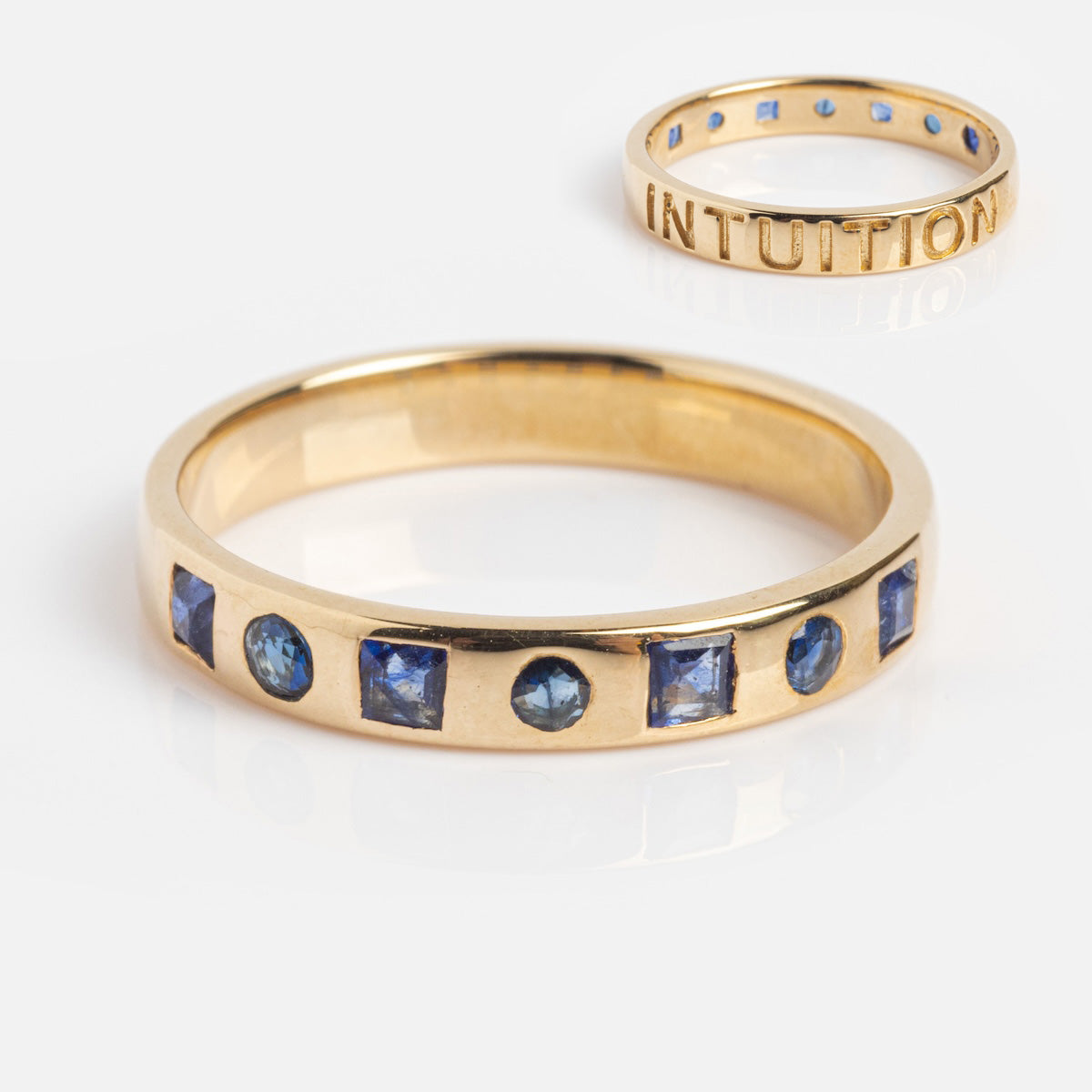 Close-up of the Solid Gold Within You Birthstone Band Sample Size 7 in 9k yellow gold with alternating round and square natural blue stones; background shows a similar ring with blue stones and INTUITION engraved on the 3.1 mm band.