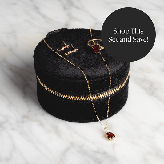 The Solid Gold 2025 January Capsule Garnet Statement Set with Free Gift—ring, necklace, and earrings with red gemstones—sits in a black velvet case with a gold zipper on marble. Shop This Set and Save!.