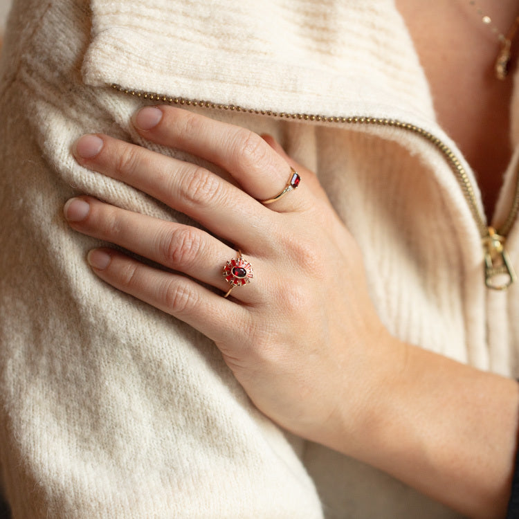 A close-up of a hand resting on an arm, showcasing the Solid Gold 2024 January Capsule Vintage Inspired Statement Ring (Sample Size 10) with garnet gemstone. The person wears a cream-colored, zippered, textured sweater.