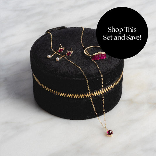 The Solid Gold 2025 July Capsule Ruby Statement Set features a ruby necklace, matching earrings, and a ring with pearl accents in a black velvet case. A “Shop This Set and Save!” overlay highlights the included free gift.