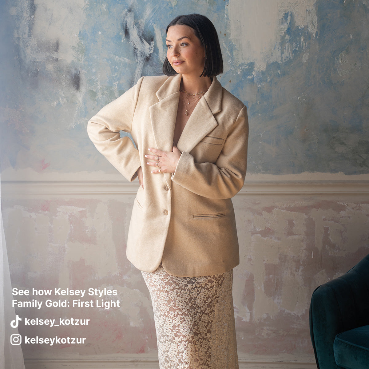 A woman in a beige blazer and lace skirt stands indoors by a painted wall, hands on her waist. She looks to the side, highlighting Solid Gold Horizon Moonstone and Opal Drop Earrings styled by Kelsey Kotzur for Family Gold: First Light.