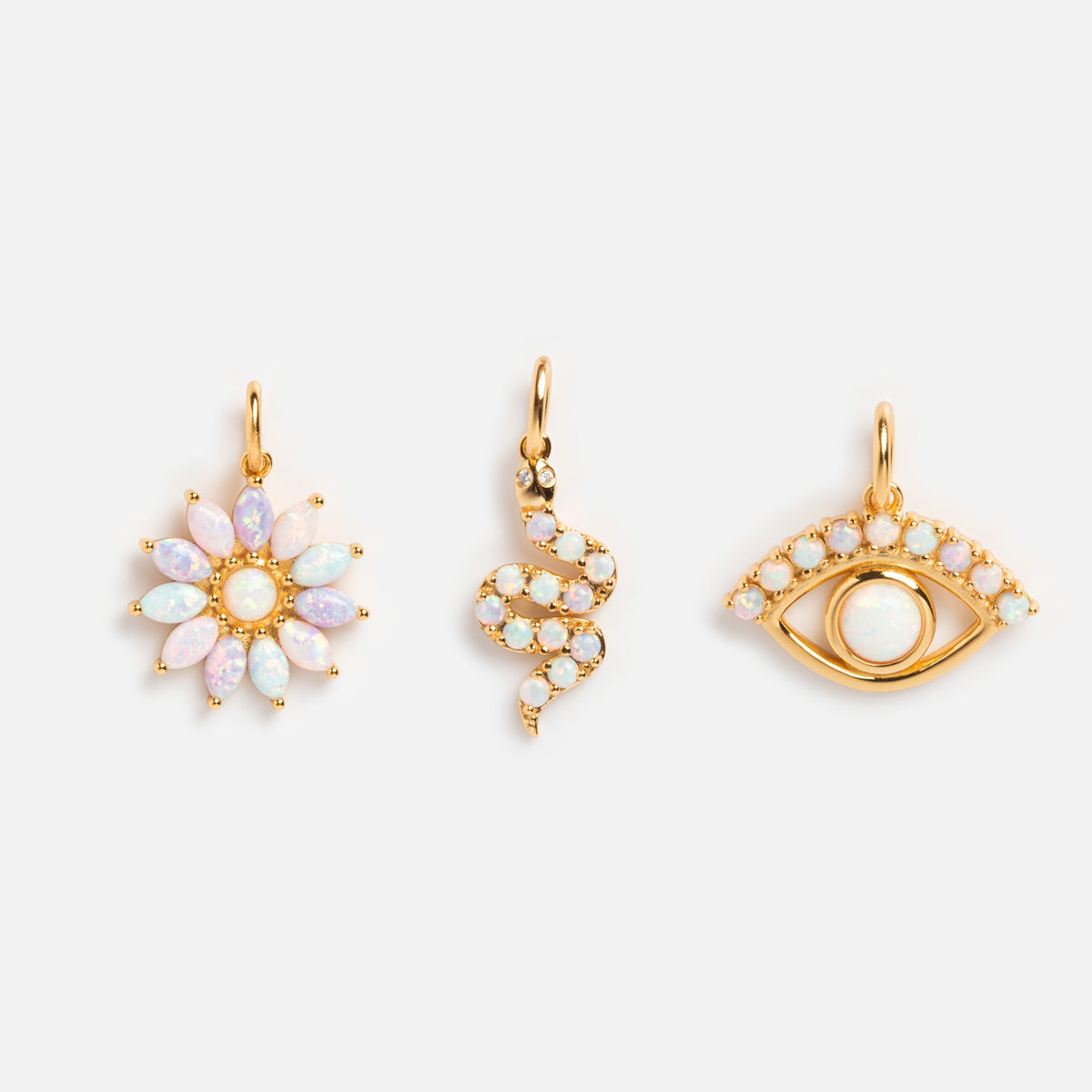 Charms by La Kaiser: Three gold pendants—a flower set with oval opals, a coiled snake with lab-created opal charms, and an eye outlined in small opals with a central stone—are showcased on a white background.