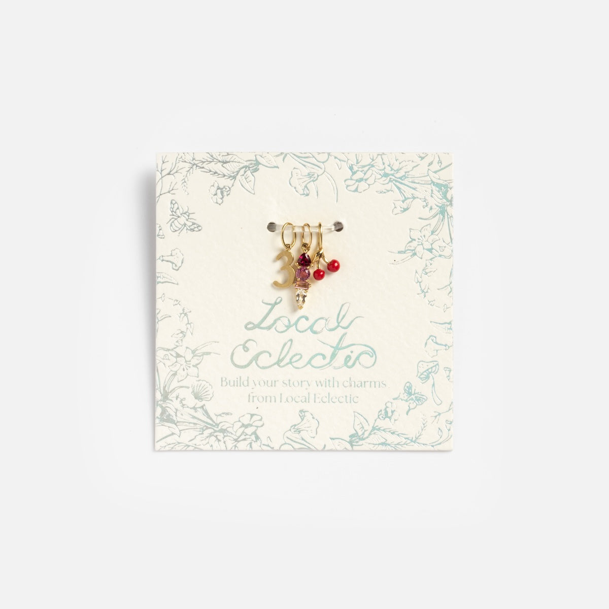 The Solid Gold Statement Charm set includes three small 9k yellow gold charms—a pearl, a blue topaz pendant, and a red beaded cluster—presented on a floral Local Eclectic card.