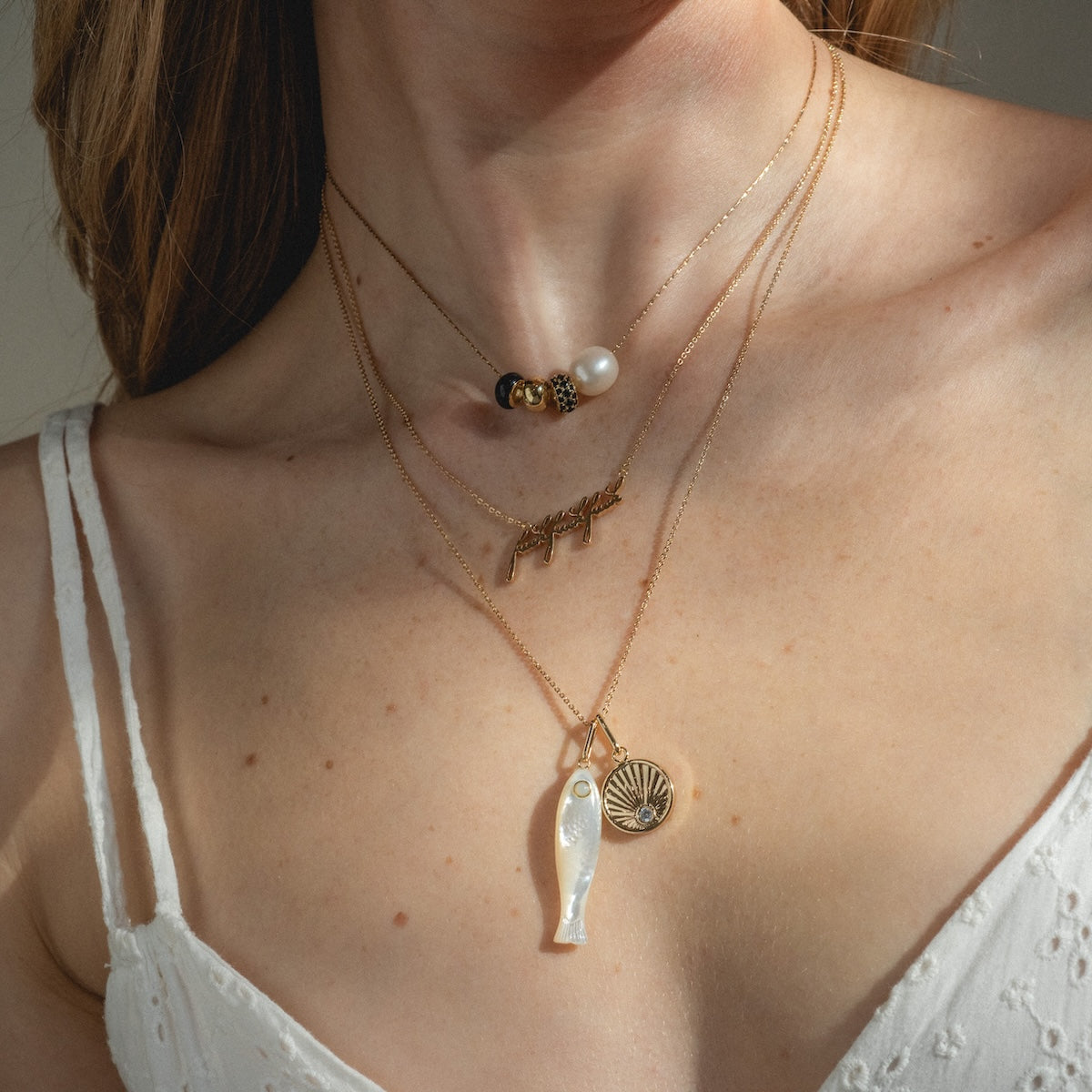 A woman wears a white, textured, sleeveless top and four layered gold Charms by Thatch necklaces featuring bead and mother of pearl charms, a word, a fish, and a circular sunburst pendant.