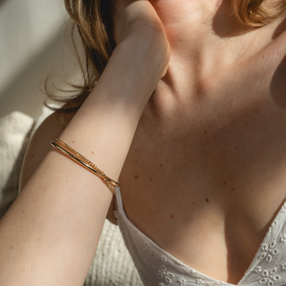 A close-up of a woman with light skin wearing a white eyelet dress and the Daniella Cuff Bracelet Sample, a delicate 14k yellow gold chain, on her wrist. Her hand rests near her neck as sunlight softly illuminates her.