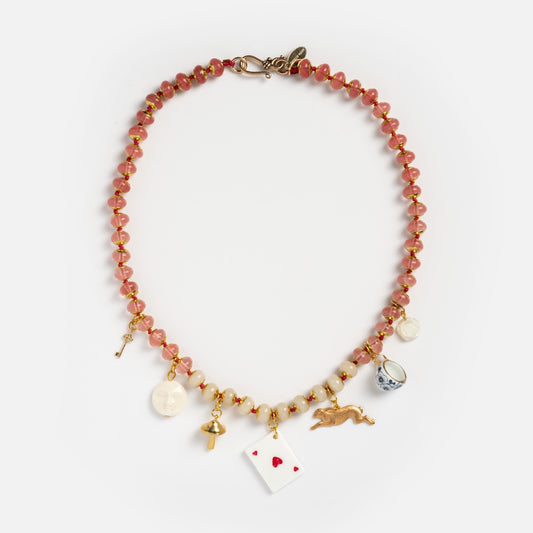 The Wonderland Necklace is a beaded necklace with alternating pink and gold beads, adorned with gold and white charms including a key, face, cross, playing card, running rabbit, teacup, and coin.