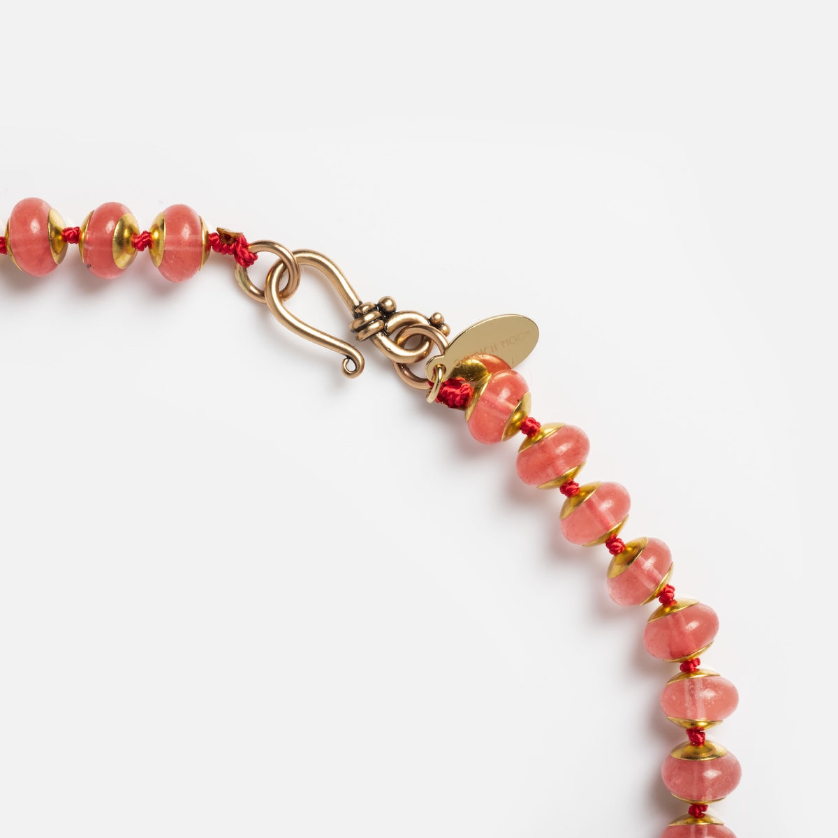 Close-up of the Wonderland Necklace featuring pink beads separated by small gold spacers, a gold hook-and-eye clasp, and an oval gold tag engraved with text, set against a white background.