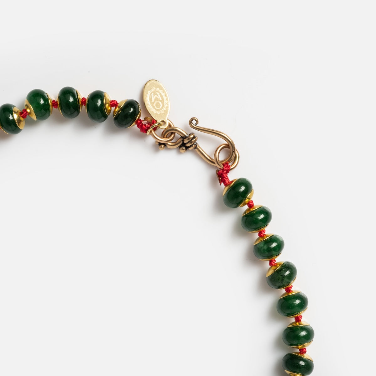 A close-up of the Zodiac Daydream Necklace, featuring dark green jade round beads with gold and red spacers, a gold hook clasp, and a gold oval tag with a decorative logo, displayed on a white background.