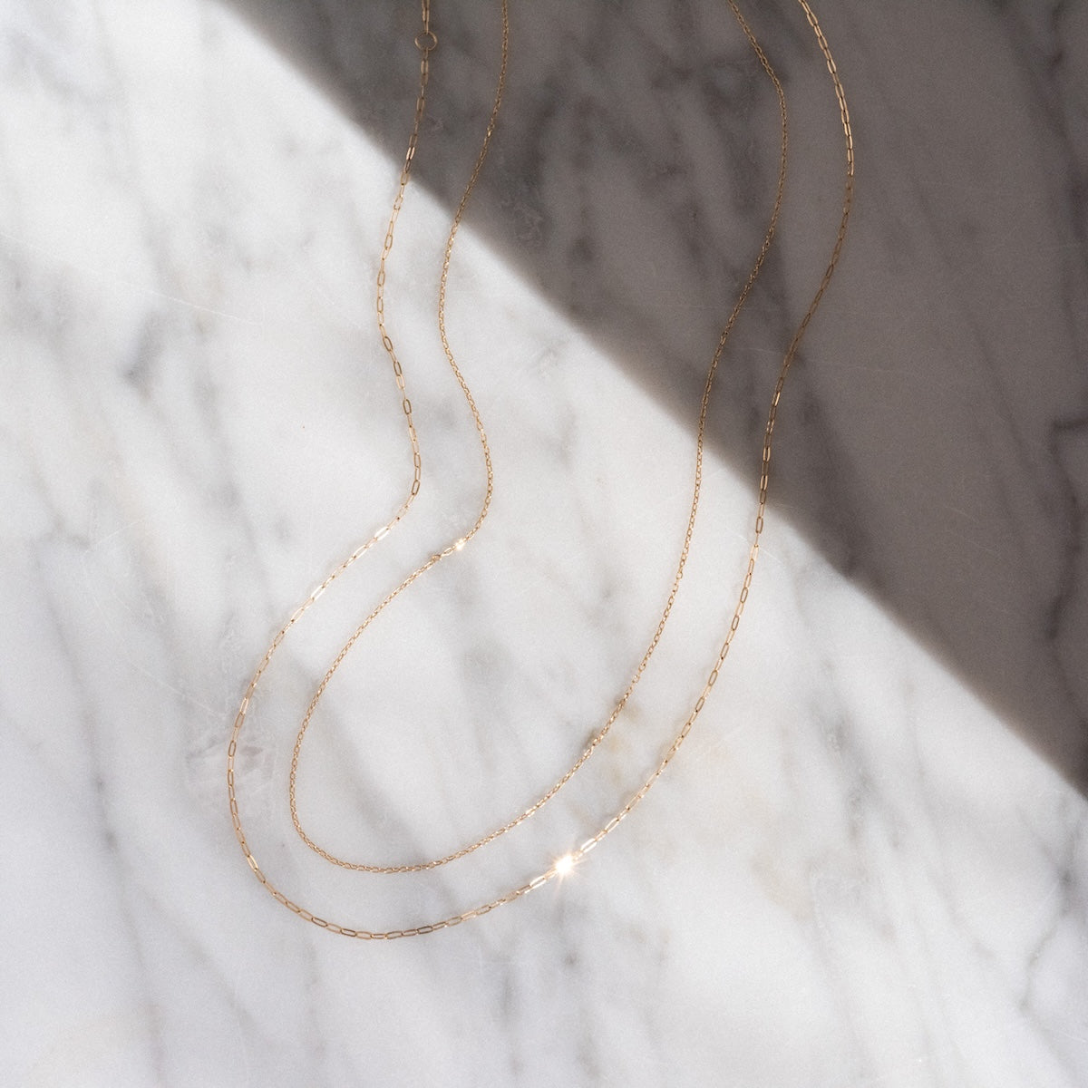 A Solid Gold Chain for Charms is elegantly looped on a white marble surface, with sunlight gently casting soft shadows across the delicate jewelry.