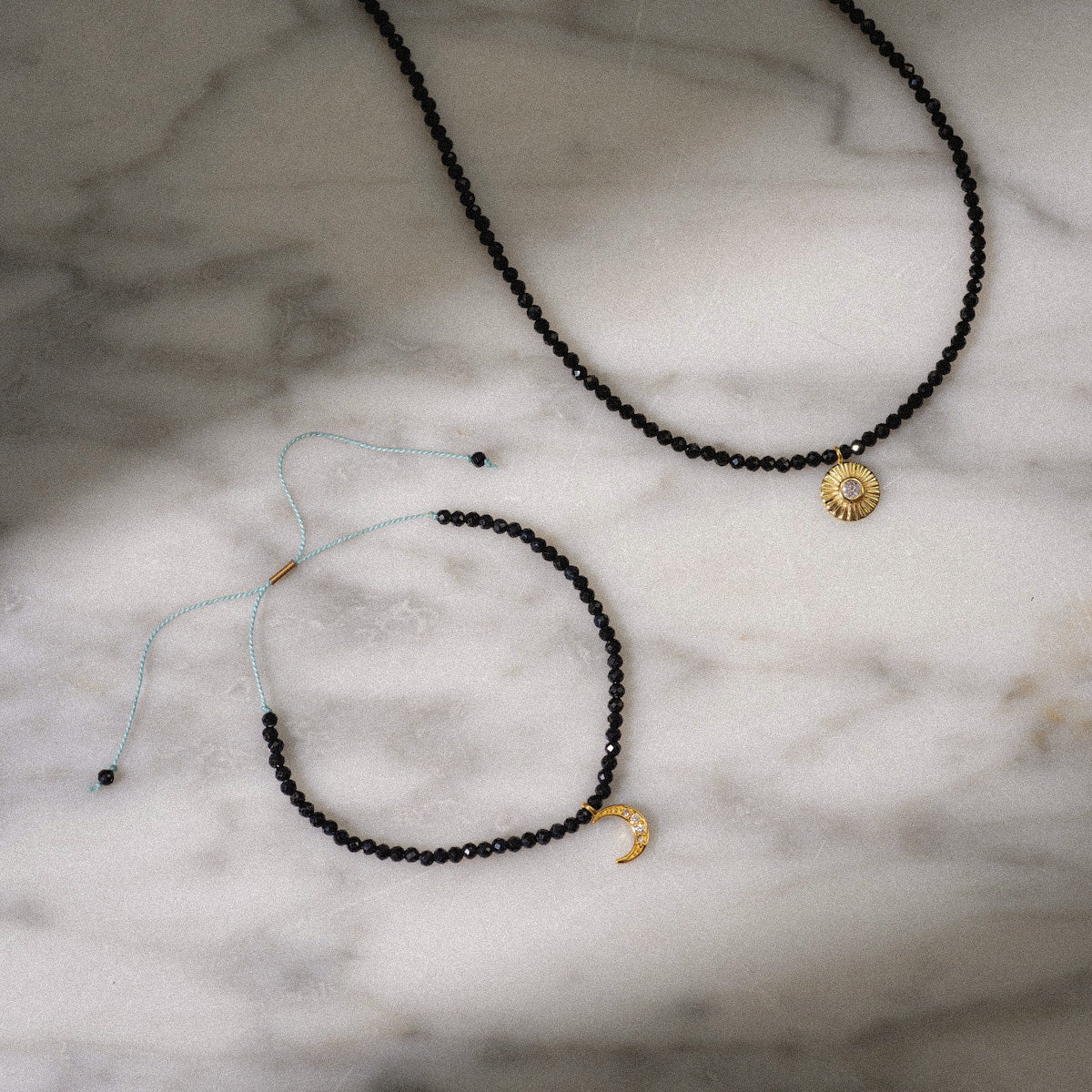 Two 10k Clarity Tiny Gemstone Necklaces rest on marble—one with a solid gold sun charm, the other adorned with spinel beads and a crescent moon charm, featuring an adjustable string closure.