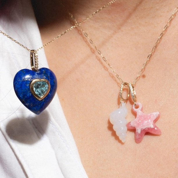 Close-up of two solid yellow gold necklaces worn over a white shirt: one with a blue heart pendant and the other featuring the 14k Pink Rhodochrosite and Diamond Sea Star Charm.